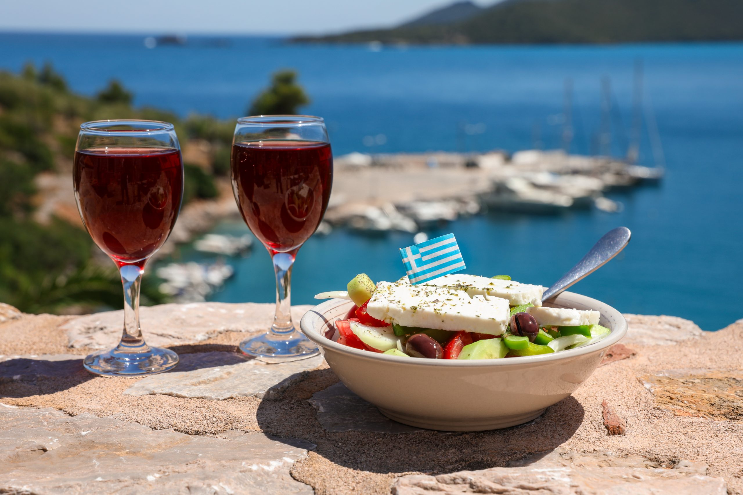 Two glasses of red wine and bowl of greek salad with greek flag on a stone terrace by the sea view, summer greek holidays concept. Horizontal. Daylight.
